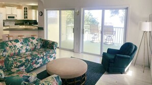 Ein Wohnzimmer mit einer Couch, einem Hocker und einem Sessel, im Hintergrund eine Küche und eine Glastür, die zu einem Außenbereich führt.