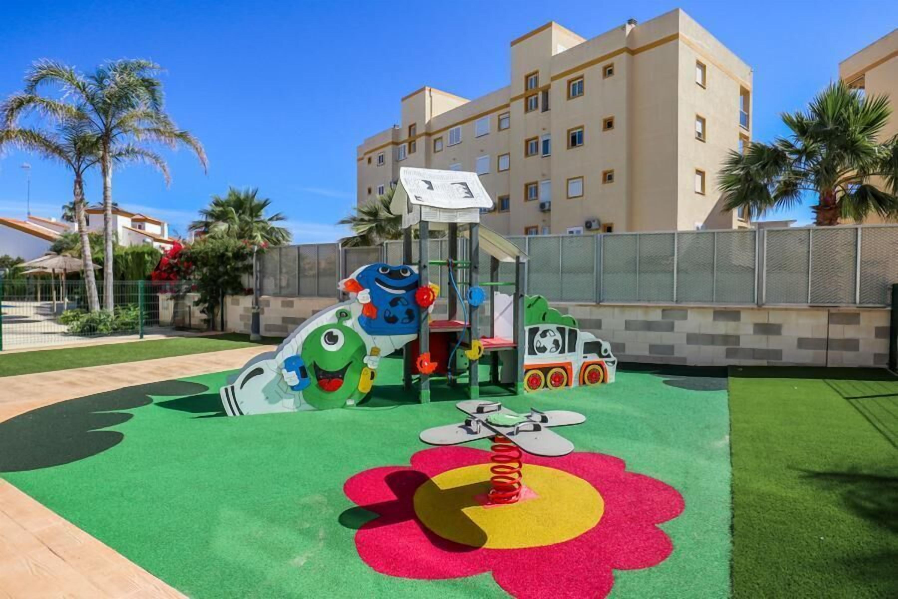 A children's play area with a slide and climbing structures, surrounded by a fence and palm trees.