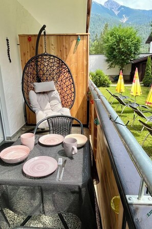 A balcony with a hanging chair, a table set with pink plates and a cup, and a view of mountains and umbrellas. - Am Birkenhain by Interhome (Seefeld in Tirol)