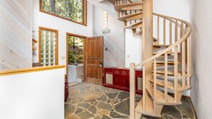 Un escalier en colimaçon en bois, situé dans un couloir doté d’un sol en pierre, de murs lambrissés et d’une grande fenêtre donnant sur un espace verdoyant.