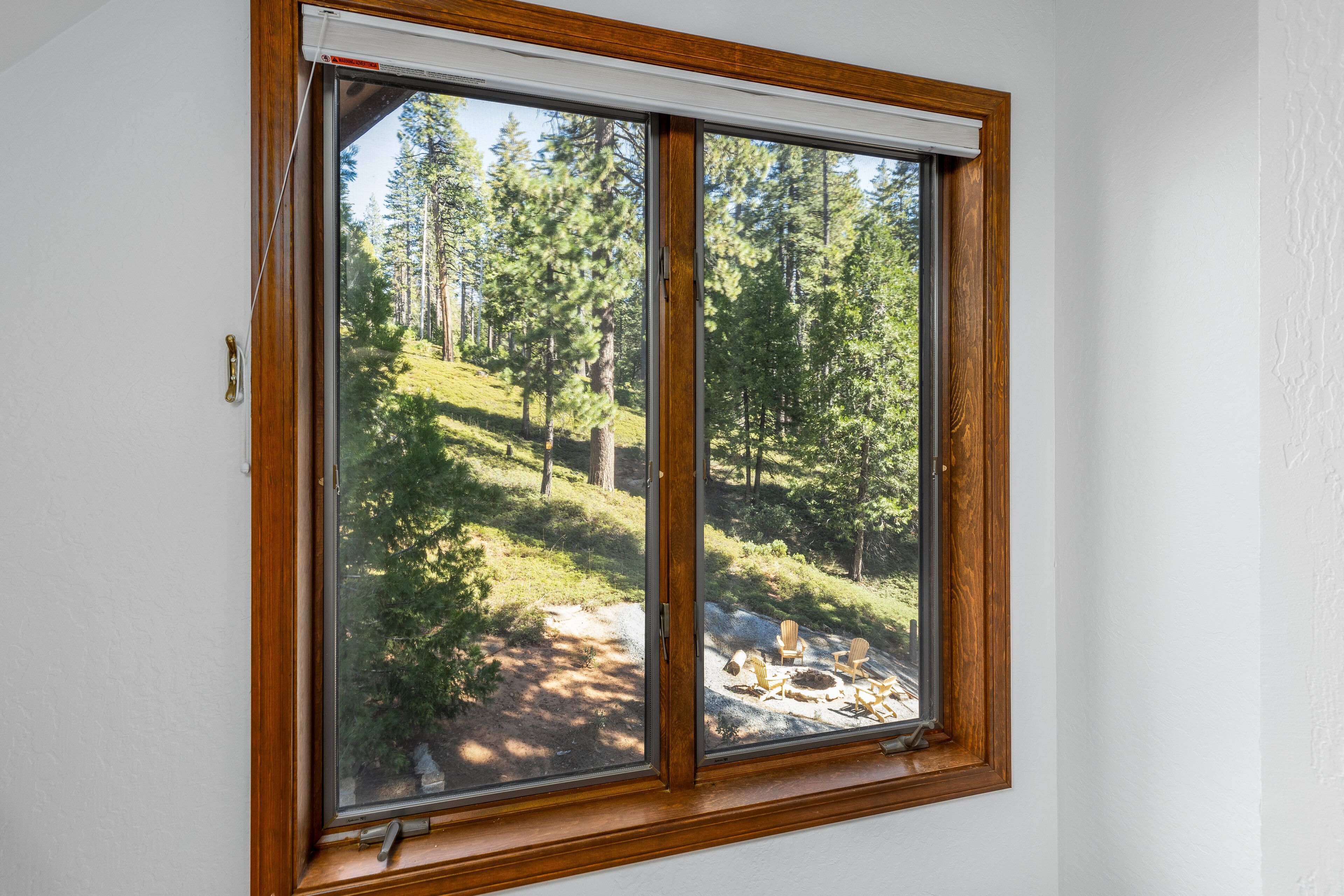 A window with a view of a forested area and a fire pit with chairs.
