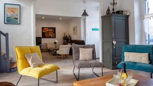 Ein modernes Wohnzimmer mit einem gelben Sessel, einem grauen Sofa und einem türkisblauen Sofa. Ein Holztisch mit einer Flasche und Gläsern steht davor. An der Wand befinden sich ein Schrank und ein Gemälde.