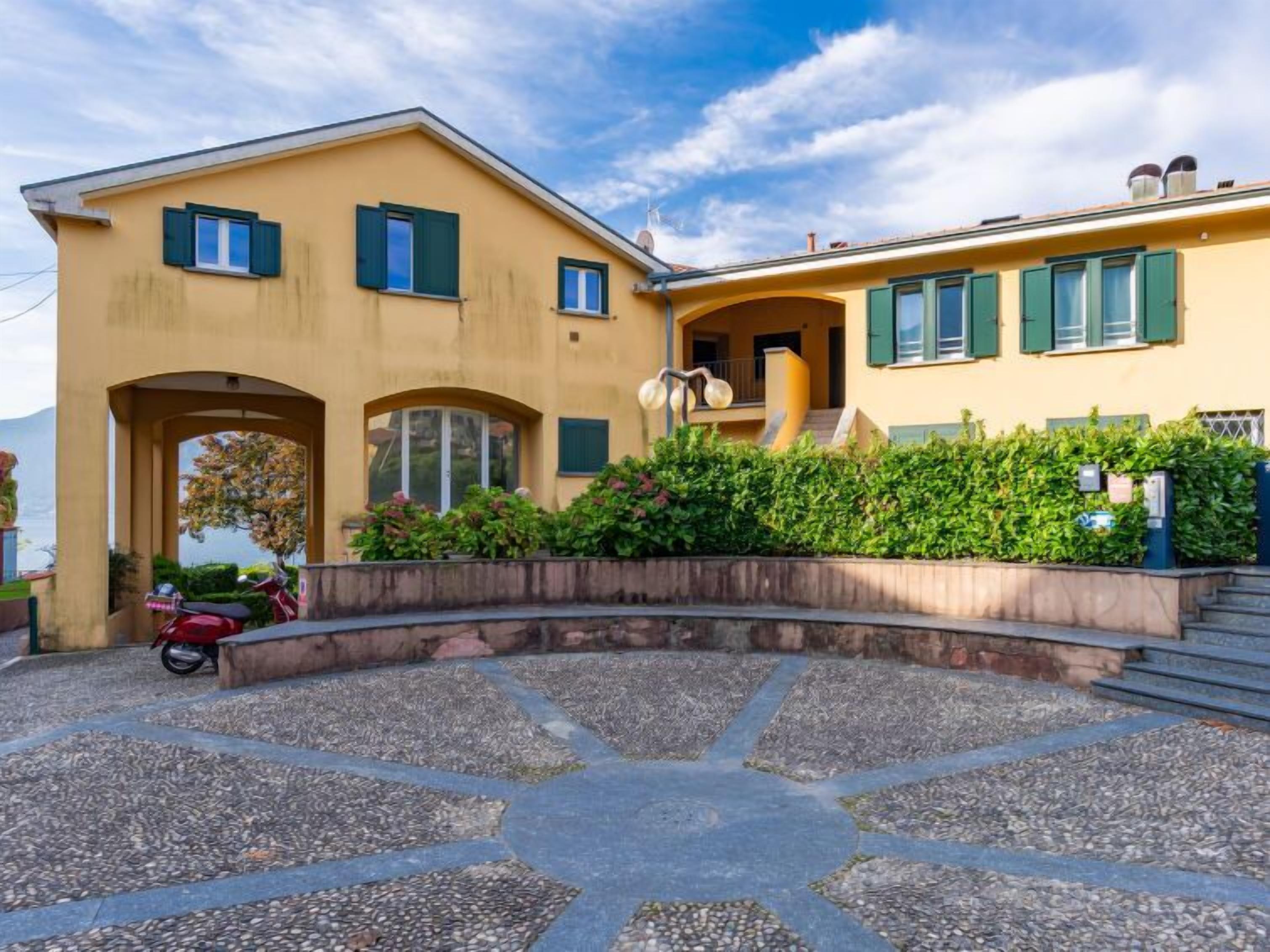 A two-story house with a yellow facade, green shutters, and a stone pathway leading to the entrance.