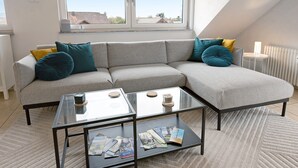 Un salon moderne avec un canapé d’angle, une table basse en verre et une vue sur l’extérieur grâce à une grande fenêtre.