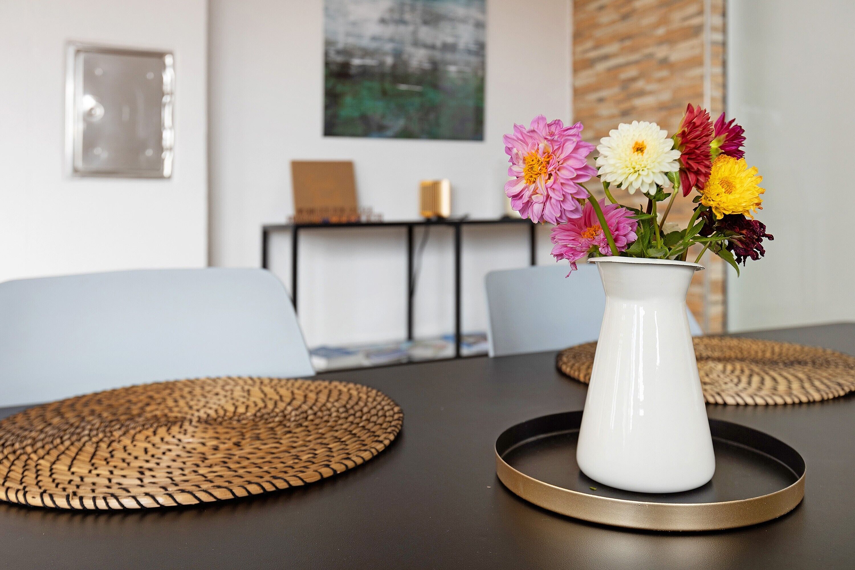 A dining table with a white vase of flowers, a laptop on a side table, and woven placemats.