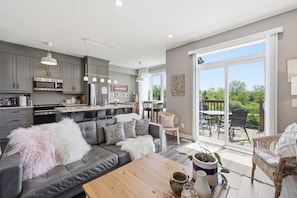 A modern kitchen with grey cabinets, a dining area with a table and chairs, a grey sofa with decorative pillows, and a large sliding glass door leading to an outdoor patio.