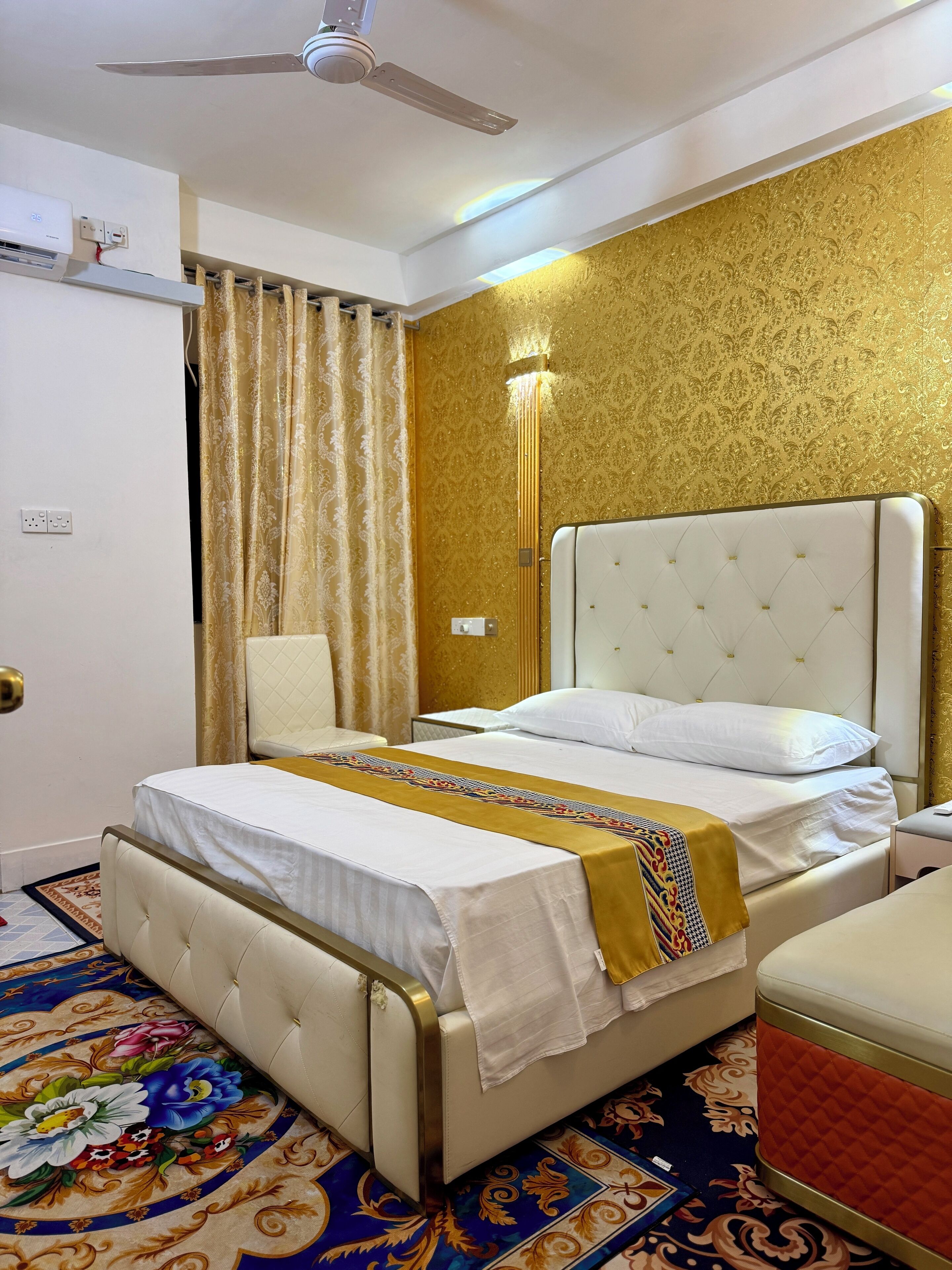 A hotel room with a bed, a chair, a bedside table, a ceiling fan, and a patterned carpet.