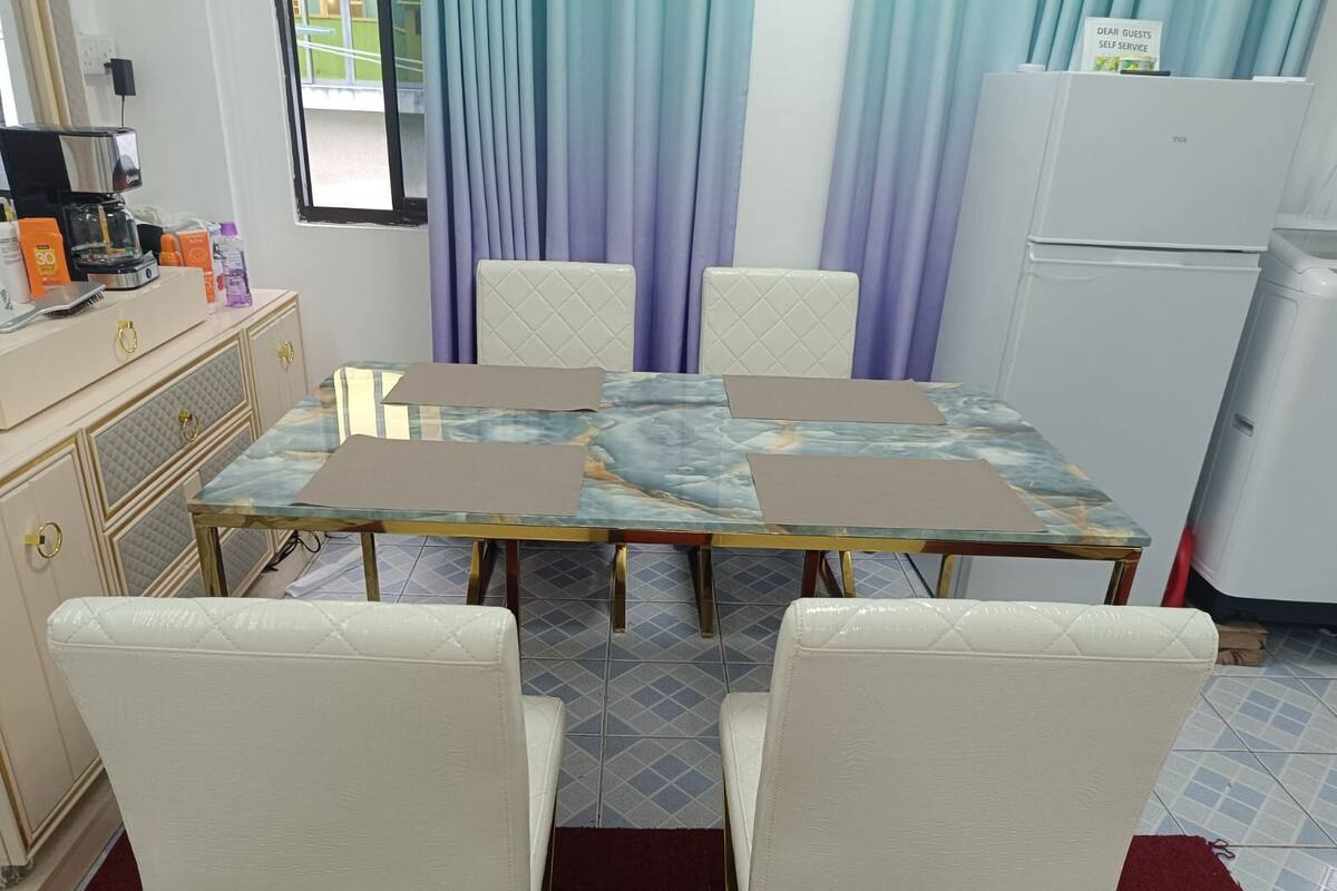 Comedor con una mesa de mármol, sillas blancas acolchadas, una cafetera y un refrigerador.