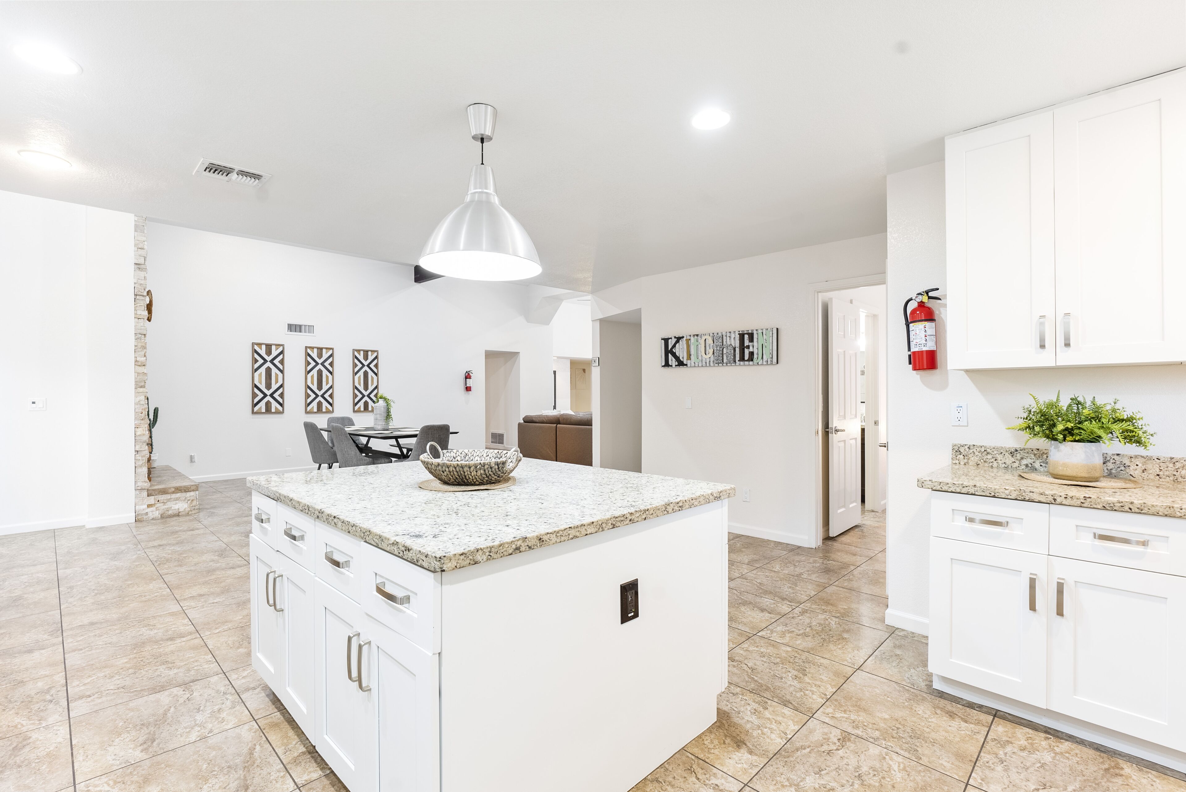 A modern kitchen with white cabinetry, a granite countertop, and a central island. There is a dining area with a table and chairs, and a kitchen island with a bowl.
