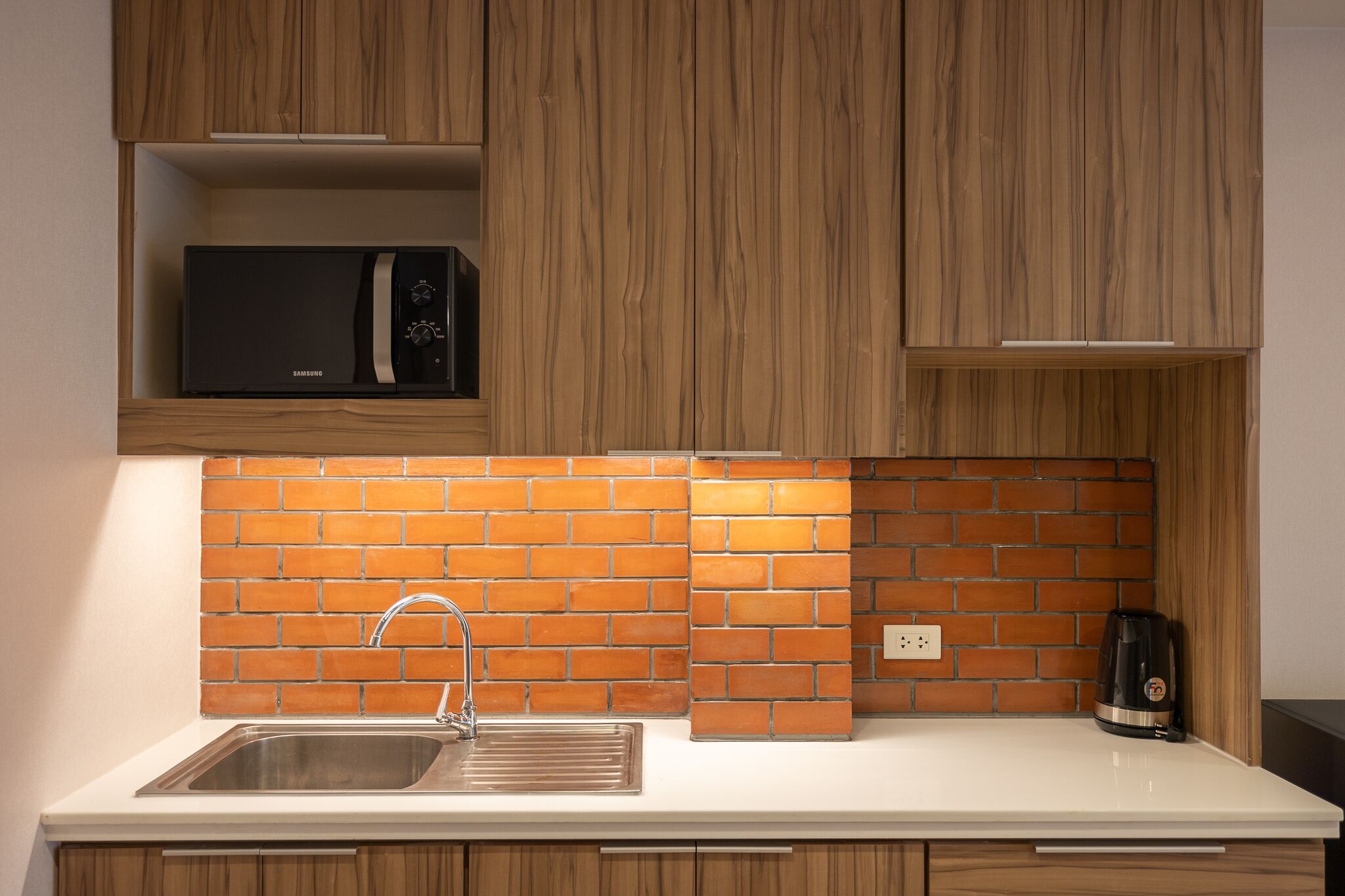 A kitchen with wooden cabinets, a brick backsplash, a stainless steel sink, a microwave, and a kettle.