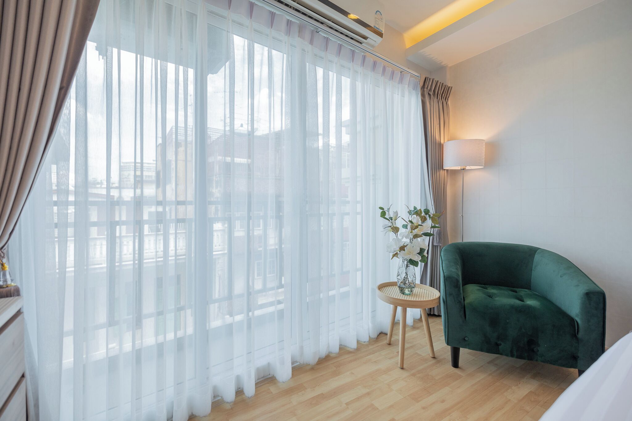 Habitación de hotel con un ventanal grande, un sillón verde, una mesita redonda con un jarrón de flores y una lámpara en la mesita auxiliar.
