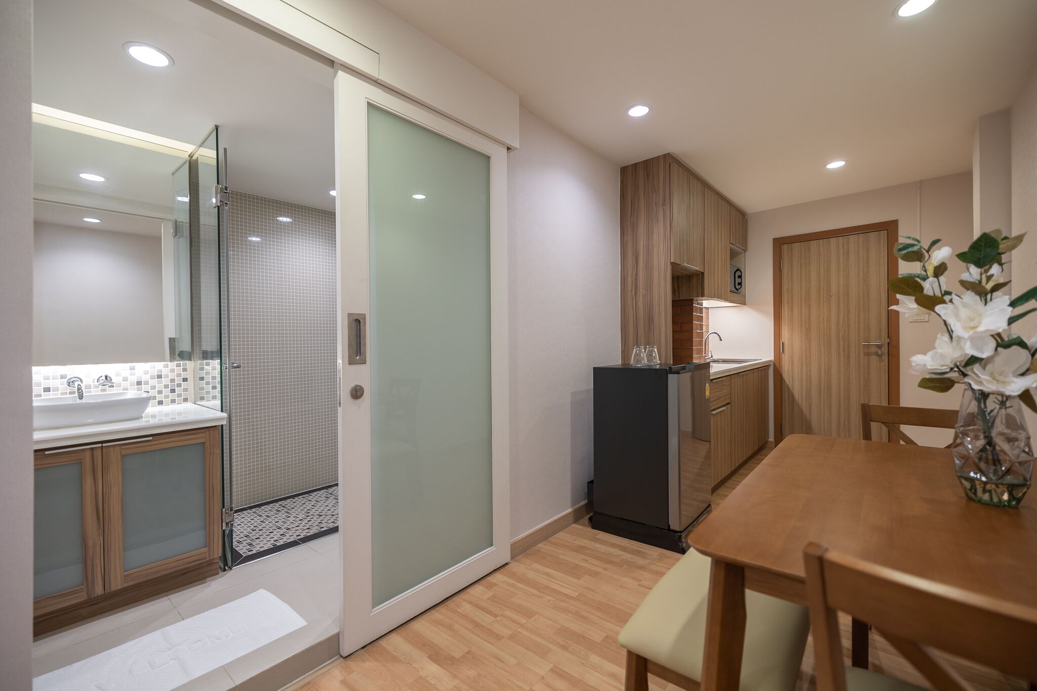 A modern kitchen with a glass door leading to a shower, wooden cabinets, and a dining table with chairs.