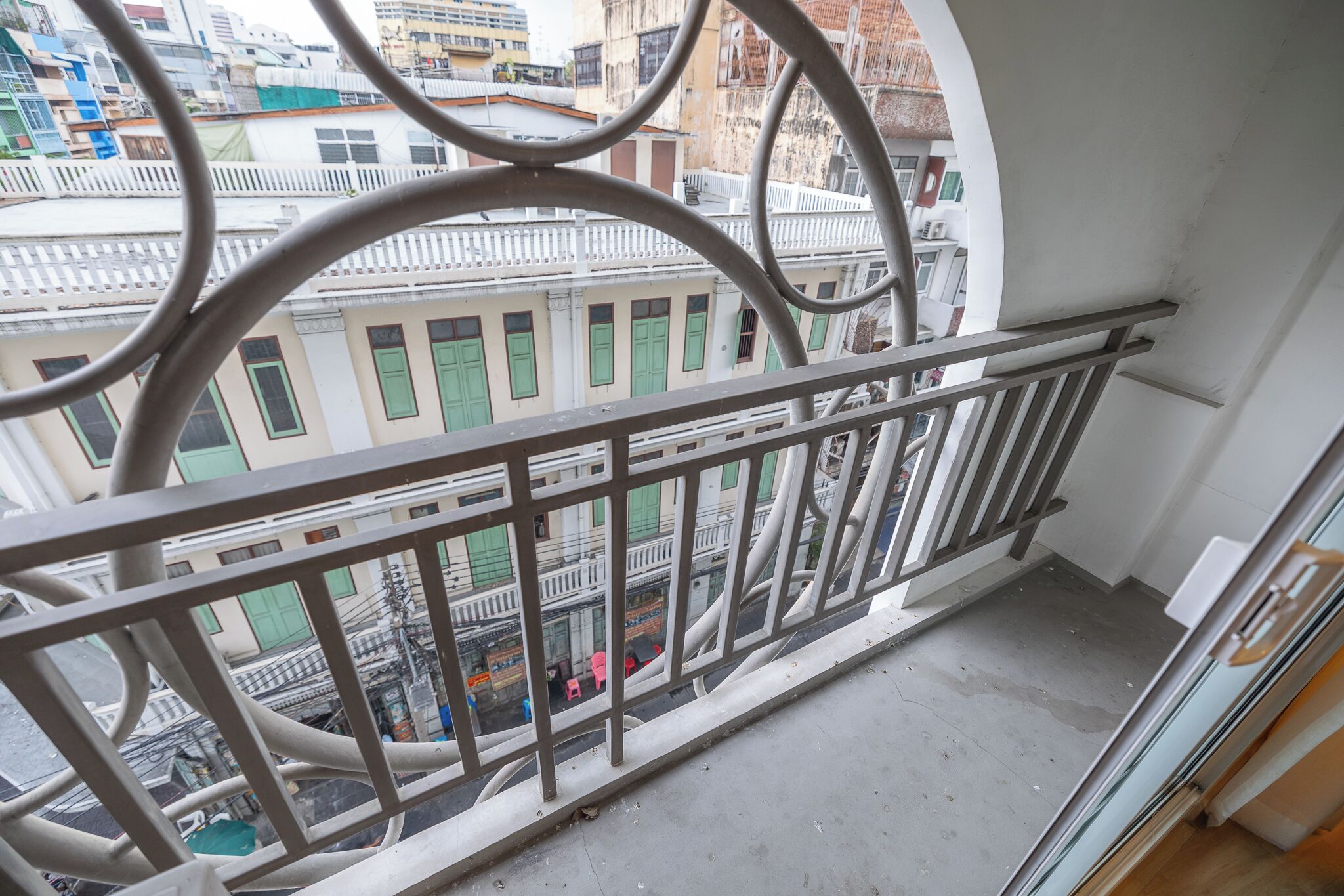A balcony with a metal railing overlooking a street with buildings and parked cars.