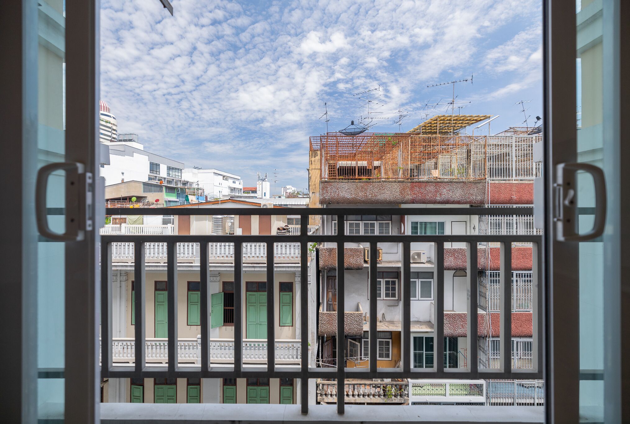 A view through a window with a metal railing, featuring multi-story buildings with balconies and a partly cloudy sky.