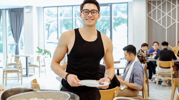 A person serving food in a well-lit dining area with large windows, wooden chairs, and tables. - RUS Hotel&Convention (Ayutthaya)