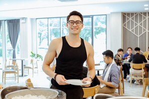 A person serving food in a well-lit dining area with large windows, wooden chairs, and tables. - RUS Hotel&Convention (Ayutthaya)