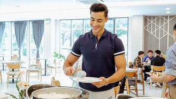 Disposición de un bufé con una persona sirviendo comida, mesas, sillas y vistas al exterior a través de grandes ventanales.
