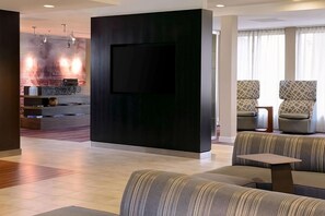 Eine moderne Hotelhalle mit einem an einer dunklen Wand montierten Flachbildfernseher, einer Sitzecke mit gemusterten Sofas und einem Empfangstisch mit Marmorplatte.