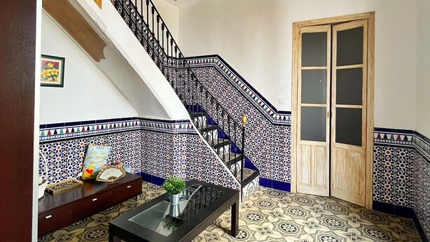 Une pièce avec un sol carrelé, un escalier dont les marches sont recouvertes de carreaux bleus et blancs, une porte en bois et une petite table sur laquelle est posée une plante.