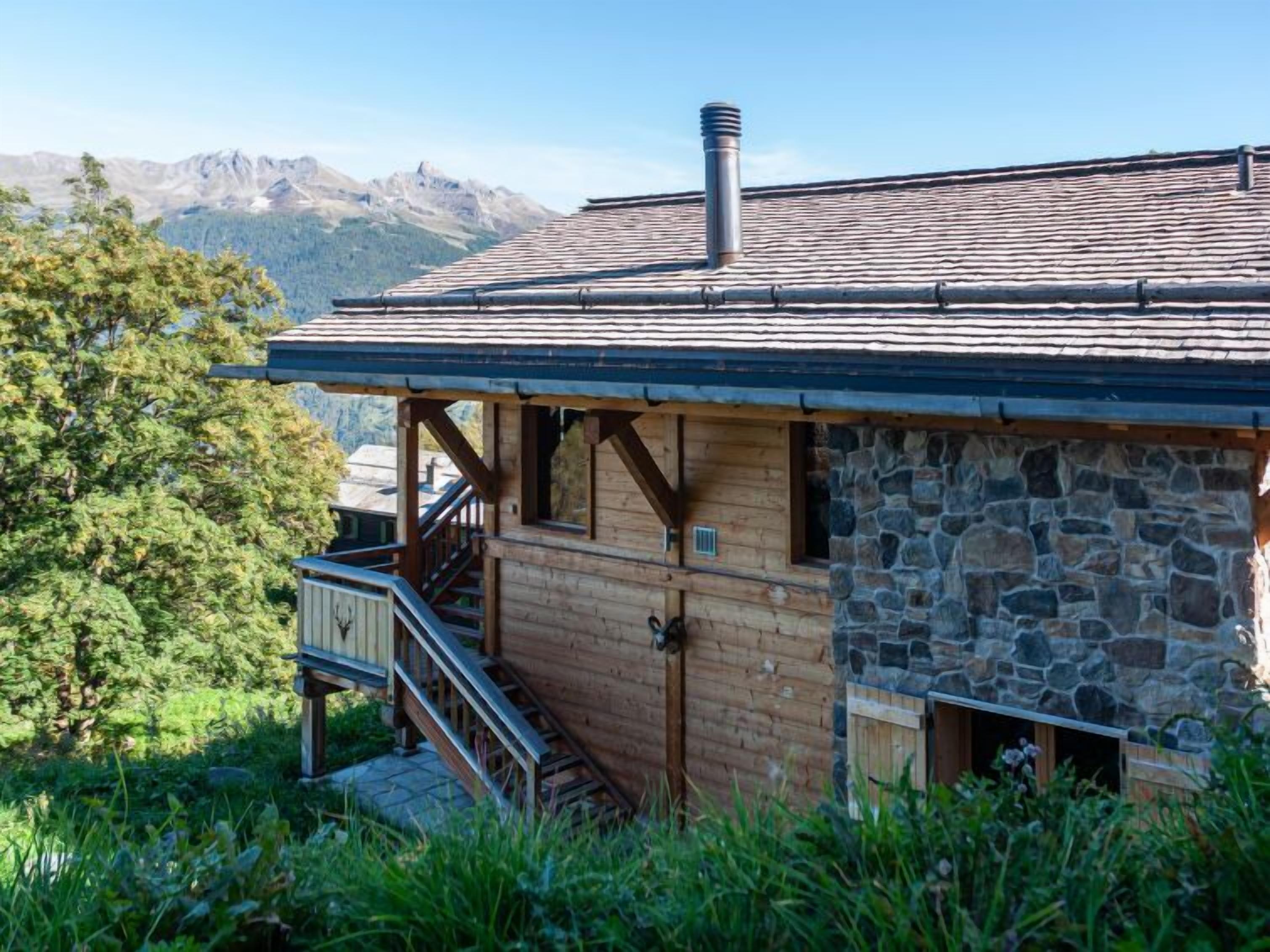 A wooden cabin with a stone exterior, a chimney, and a balcony overlooking a mountainous landscape.