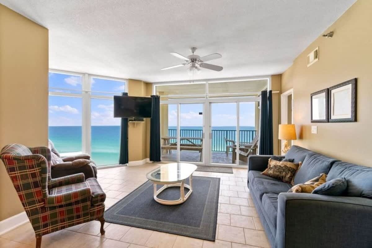 A living room with a view of the ocean, a ceiling fan, a flat-screen TV, and a sofa with cushions.