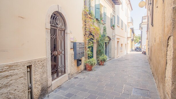 A narrow cobblestone street lined with buildings featuring arched windows and potted plants. - Old Town Alley - Italian Homing (Desenzano del Garda)