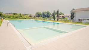 A rectangular swimming pool with a tiled deck, surrounded by greenery and buildings in the background.