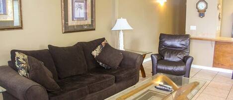 A living room with a brown sofa, a glass coffee table, a leather armchair, and framed pictures on the wall.