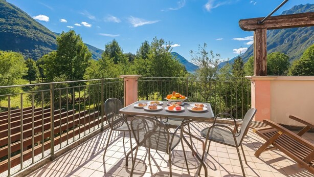 A balcony with a table set for dining, chairs, and a view of mountains. - Porto Letizia Begonia 7, Porlezza, Italy (Porlezza)