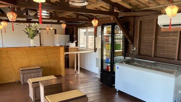 A wooden interior with a reception desk, a refrigerator stocked with beverages, and hanging lanterns. - Pension Shimakaze (Motobu)