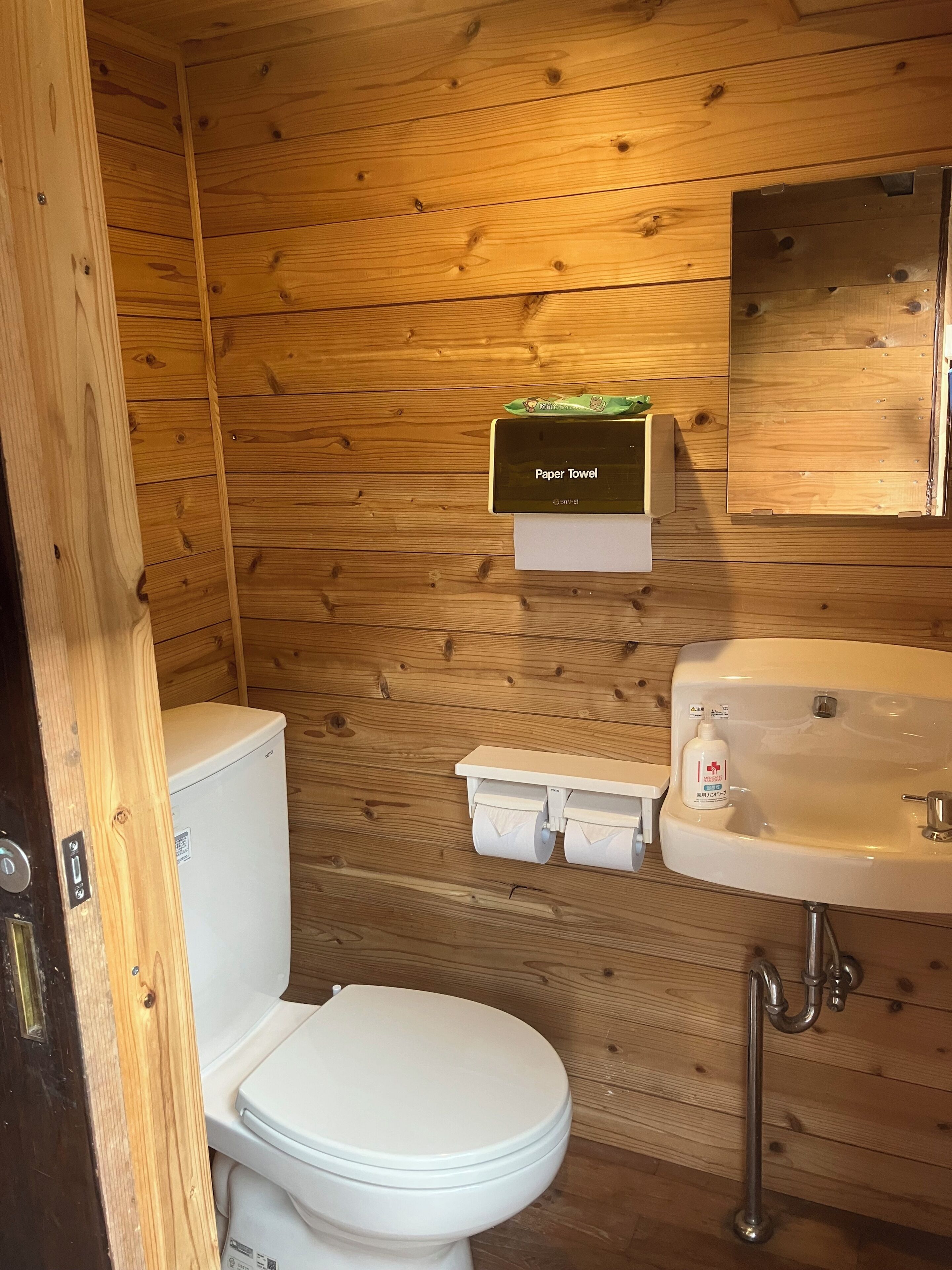 A wooden-paneled bathroom with a toilet, paper towel dispenser, and sink.