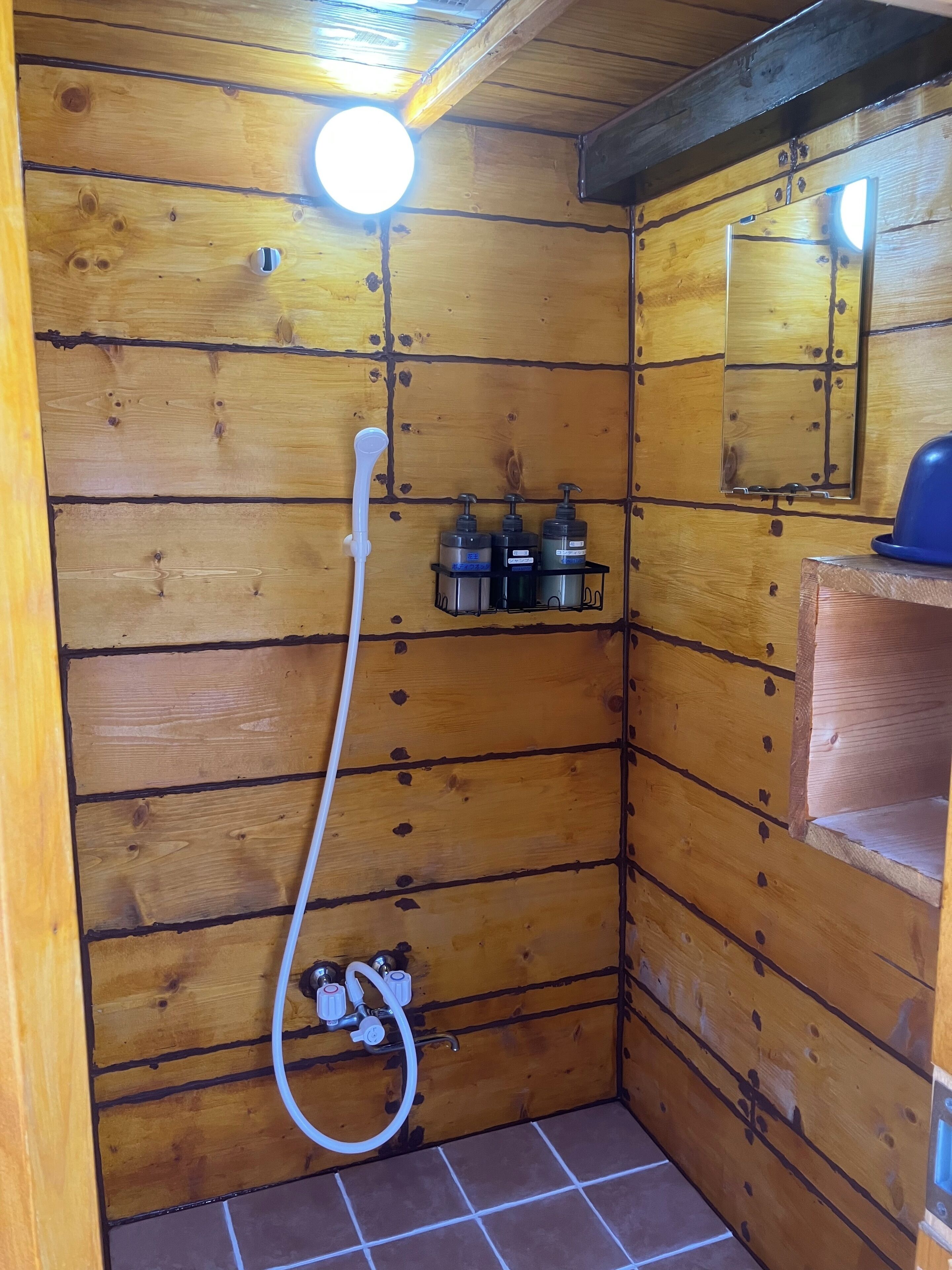 A wooden shower stall with a handheld showerhead, a wall-mounted mirror, and a shelf with toiletries.