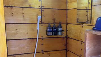 A wooden shower stall with a handheld showerhead, a wall-mounted mirror, and a shelf with toiletries.