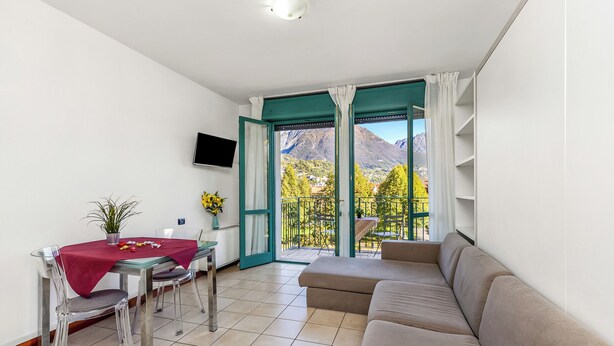 A modern living room with a sofa, dining table, and chairs. A TV is mounted on the wall, and a balcony with a view of mountains is visible through the open door. - Porto Letizia S1, Porlezza, Italy (Porlezza)