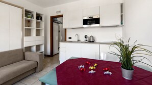 A modern kitchen with white cabinets, a built-in oven, and a coffee maker. There is a red table with a potted plant and wrapped chocolates. - Porto Letizia S1, Porlezza, Italy (Porlezza)