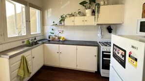 A kitchen with white cabinets, a stove, and a refrigerator. There are plants on the shelves and a window with curtains. - Cosy Flat metres from Rottnest Ferry (Fremantle)