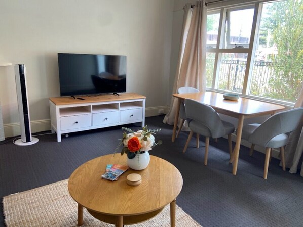 A room with a TV, a wooden table with a vase of flowers, a desk with chairs, and a window with a view of trees.