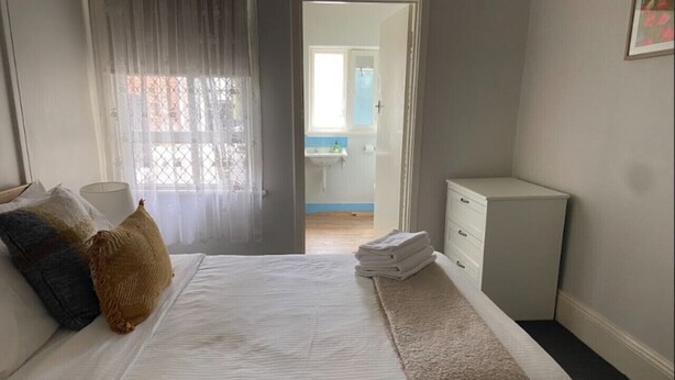 A neatly made bed with a white comforter and a beige throw blanket, a small bedside table with a lamp, and a view of a bathroom through an open door. - Brilliant Apt by Rottnest Ferry and Fremantle Oval (Fremantle)