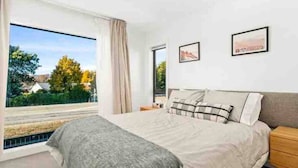 A bedroom with a large bed, a nightstand, and a window with a view of trees and houses. - Wanaka Town House - Single Malt (Albert Town)