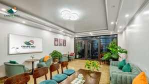 En moderne hotellobby med en træreception, teal stole og grønne sofaer. Der er plantede planter og et stort vindue med udsigt ud.