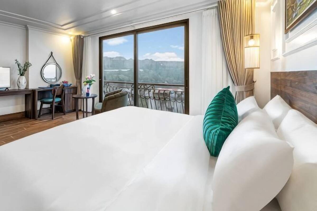 A hotel room with a large bed, a desk with a chair, a small table with a vase, and a balcony with a view of the mountains.