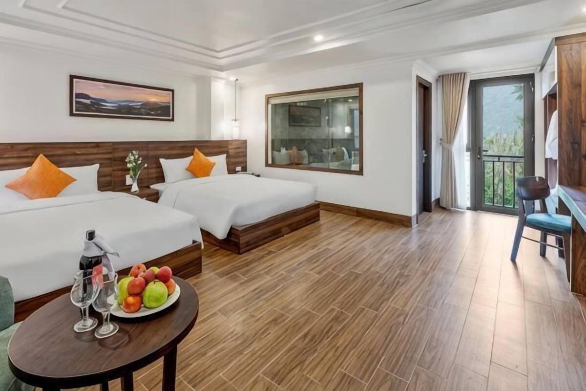 A hotel room with two beds, a balcony, a dining table with fruit and drinks, and a view of the outdoors.