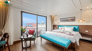 Et hotelværelse med en stor seng, en træbænk, et lille bord med en vase blomster, en stol, samt en balkon med udsigt til bygninger og et vandtårn.