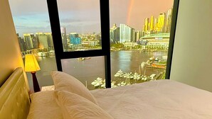 A hotel room with a bed, a lamp, and a view of a cityscape and a rainbow through large windows. - Ensuite Double Room with Private Bathroom and River View (Docklands)