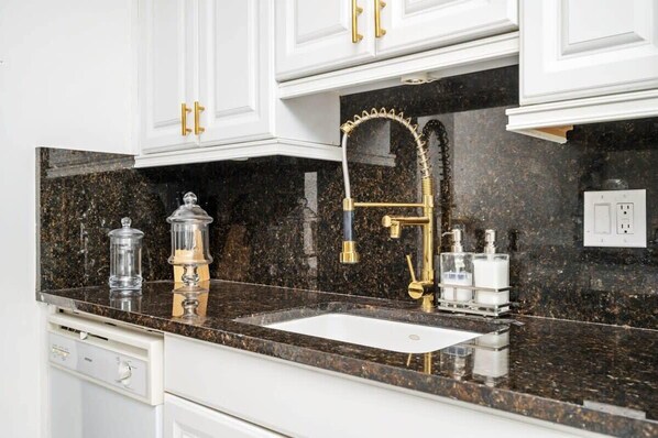Una cocina con gabinetes blancos, encimera de granito, grifo dorado y pileta blanca.