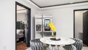 Een moderne eethoek met een marmeren tafel en gestoffeerde stoelen, een koelkast met afbeelding en een bloemstuk met gele bloemen.