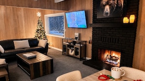 A cozy living room with a fireplace, a Christmas tree, a TV, and a coffee table with mugs and marshmallows. - SlopeSide • Hot Tub • Arcade • Deck w BBQ+fire pit (Big Bear Lake)