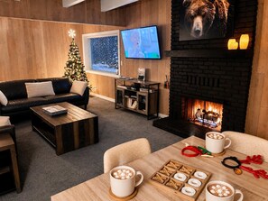 A cozy living room with a fireplace, a Christmas tree, a TV, and a coffee table with mugs and marshmallows. - SlopeSide • Hot Tub • Arcade • Deck w BBQ+fire pit (Big Bear Lake)