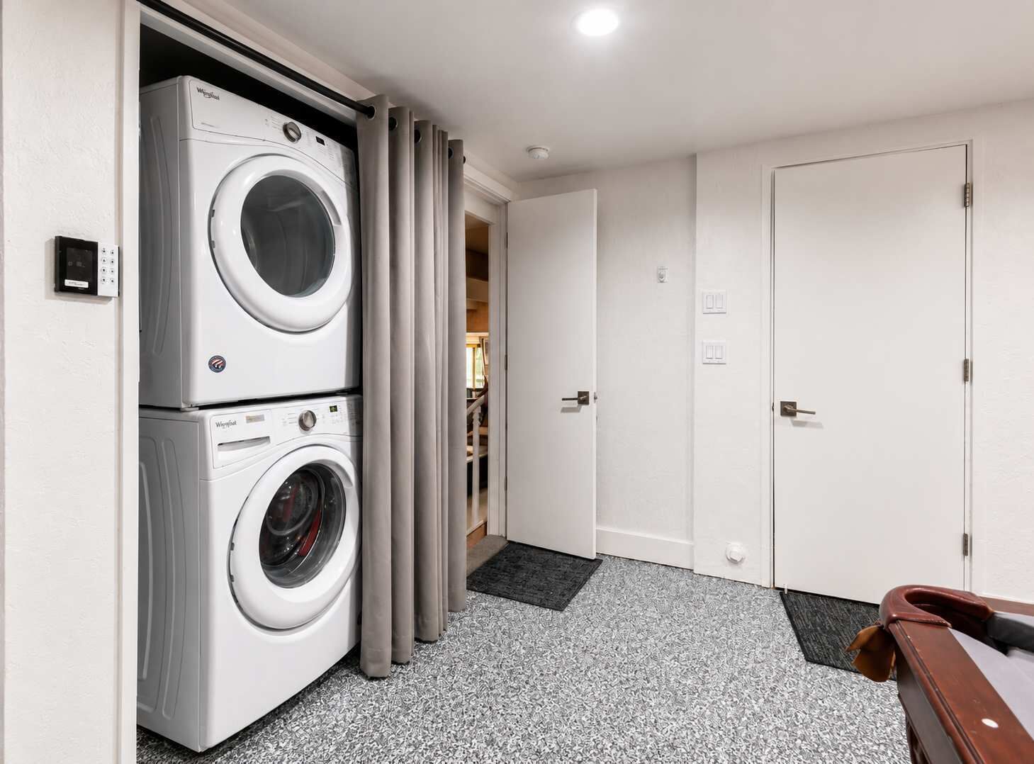Un lavadero con lavadoras y secadoras apiladas, una puerta que da a otra habitación y una pequeña mesa con una silla.