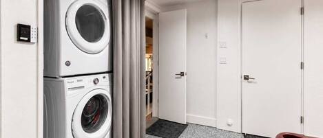 Un lavadero con lavadoras y secadoras apiladas, una puerta que da a otra habitación y una pequeña mesa con una silla.